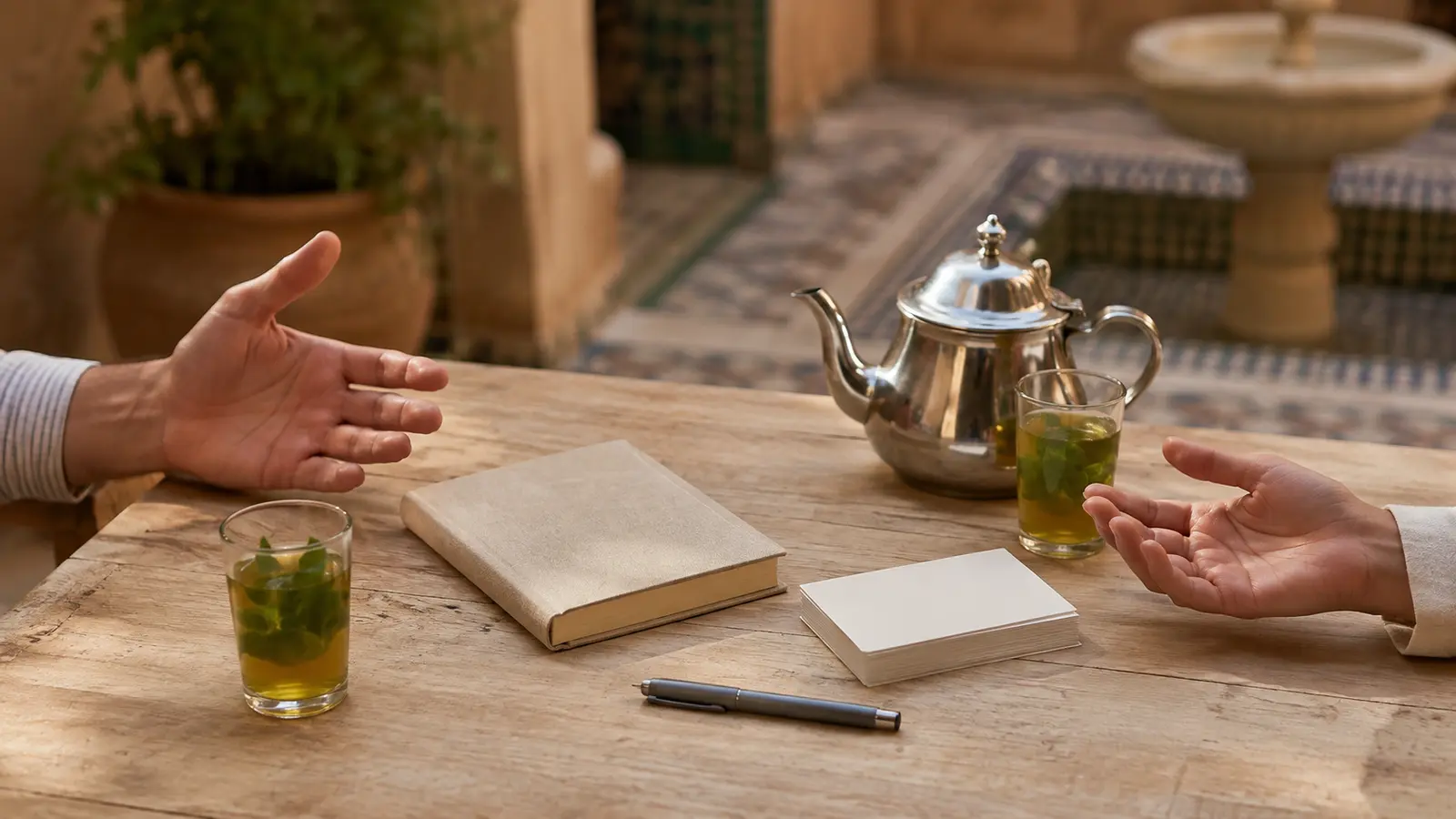 Two travelers gesture over a closed notebook, blank cards, pen and Moroccan tea